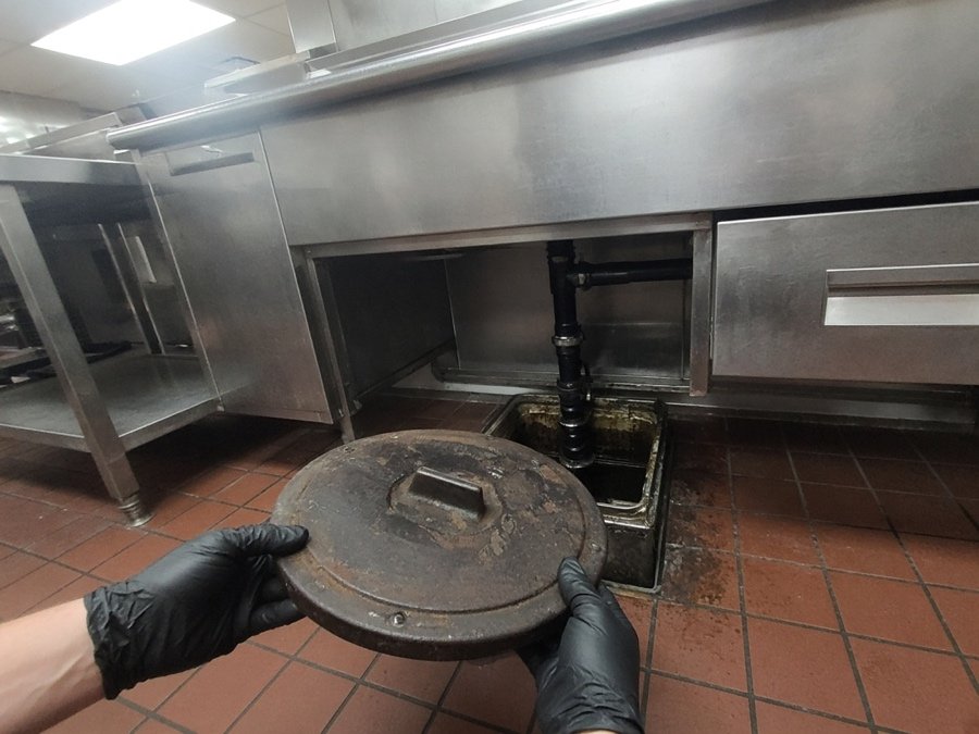 Gloved hands lifting a cast-iron grease trap lid in a commercial kitchen — grease trap cleaning in Fullerton, CA